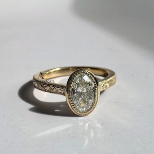 May include: A gold ring with an oval-shaped, clear gemstone. The ring has a detailed, engraved band. The gemstone is set in a bezel setting. The ring is displayed on a white surface.