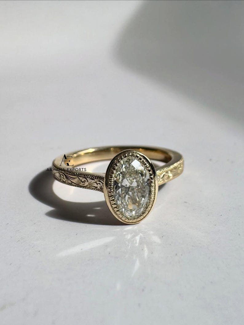 May include: A gold ring with an oval-shaped, clear gemstone. The ring has a detailed, engraved band. The gemstone is set in a bezel setting. The ring is displayed on a white surface.