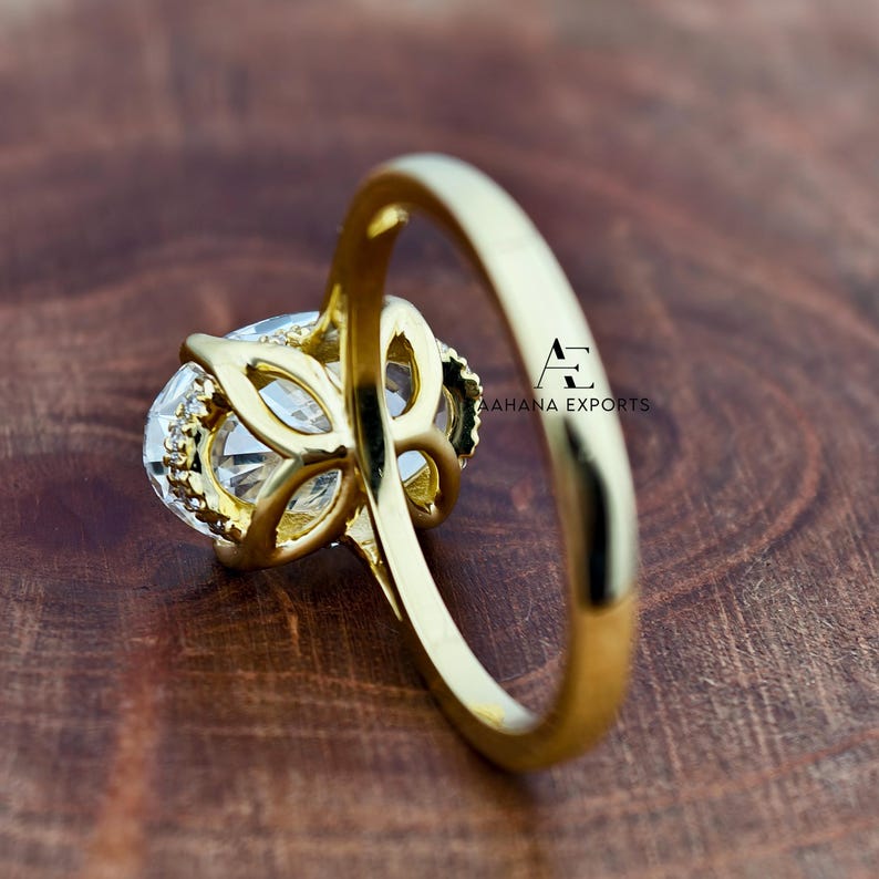 May include: A gold ring with a large, clear, oval-cut gemstone. The ring has a detailed setting with smaller accent stones. The band is smooth and polished. The ring is set against a wooden surface. The text "AAHANA EXPORTS" is visible.