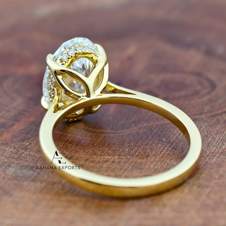 May include: A gold engagement ring with a large, clear gemstone and smaller accent stones. The ring has a detailed setting and a smooth, polished band. The logo "AAHANA EXPORTS" is visible on the band.