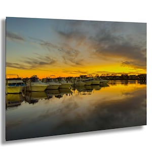 Puede incluir: Fotografía escénica de un puerto deportivo al atardecer. Varios barcos están atracados, reflejándose en el agua en calma. El cielo es una vibrante mezcla de naranja, amarillo y gris, creando una escena pacífica y pintoresca.