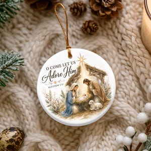 Puede incluir: Un adorno redondo de cer&aacute;mica blanca con una ilustraci&oacute;n de la escena de la Natividad con Mar&iacute;a, Jos&eacute; y el ni&ntilde;o Jes&uacute;s en un pesebre. El texto dice O COME LET US Adore Him y MATTHEW 2:11. Un cord&oacute;n dorado est&aacute; unido a la parte superior.