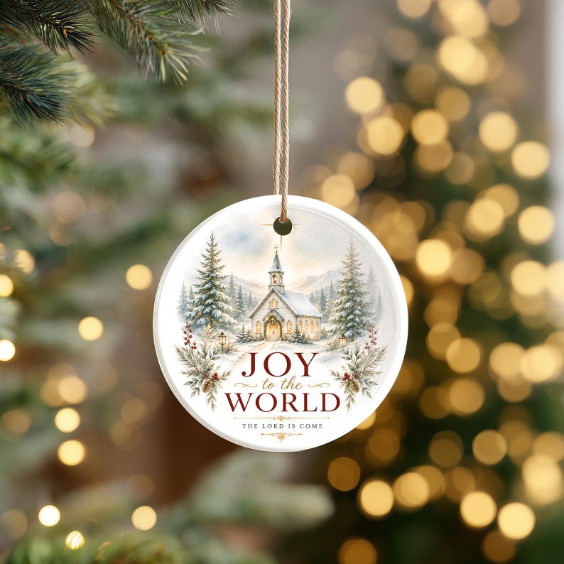 Puede incluir: Un adorno redondo de cer&aacute;mica blanca que cuelga de un cordel color canela. El frente presenta una ilustraci&oacute;n de una iglesia cubierta de nieve rodeada de pinos. El texto dice JOY to the WORLD THE LORD IS COME en letras rojas y negras.