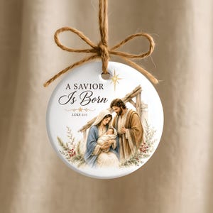 Puede incluir: Un adorno redondo de cer&aacute;mica blanca con una ilustraci&oacute;n de la natividad que muestra a Mar&iacute;a, Jos&eacute; y el ni&ntilde;o Jes&uacute;s. El texto dice A SAVIOR Is Born y LUKE 2:11. Una estrella dorada corona las figuras, colgado de un lazo de cuerda marr&oacute;n.