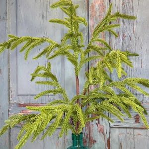 May include: A bright green artificial pine branch with needle-like foliage and a brown stem, placed inside a translucent teal glass bottle with a diamond-patterned texture. A roll of plaid ribbon with red, white, and green colors sits nearby.