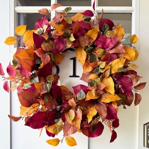 May include: A circular wreath composed of artificial autumn leaves in shades of deep burgundy, burnt orange, golden yellow, and muted green. The foliage is densely arranged, creating a full, rounded shape that displays a variety of leaf textures.