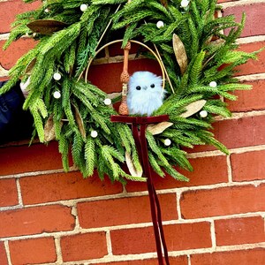May include: A decorative wreath featuring green pine needles, white berries, and gold leaves. A gold metal hoop is centered within the wreath, holding a brown wooden wand and a small, fluffy white owl figurine. A long, dark red velvet ribbon hangs down.