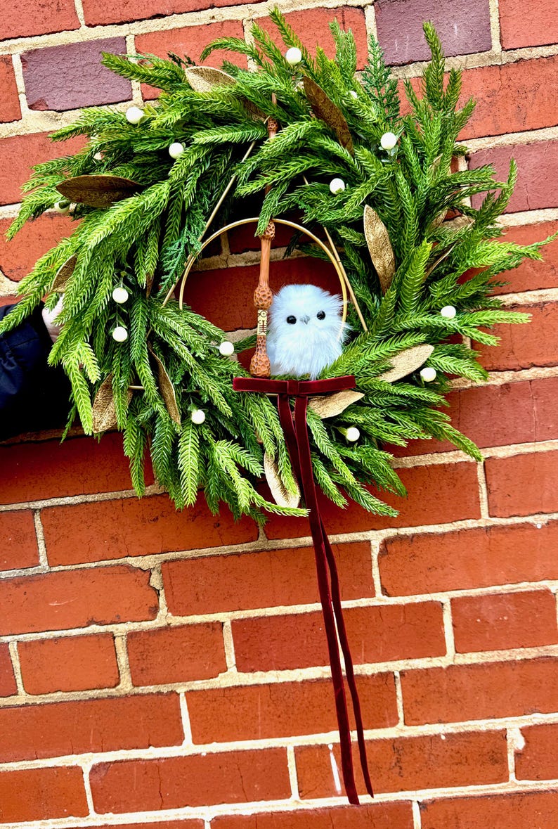 May include: A decorative wreath featuring green pine needles, white berries, and gold leaves. A gold metal hoop is centered within the wreath, holding a brown wooden wand and a small, fluffy white owl figurine. A long, dark red velvet ribbon hangs down.