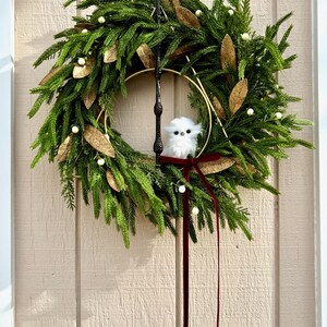 May include: A decorative wreath featuring green pine-like foliage, gold-toned leaves, and small white berries. A gold metal ring and a dark vertical rod are integrated into the design. A small, fluffy white owl figure sits inside, accented by a long dark red ribbon.