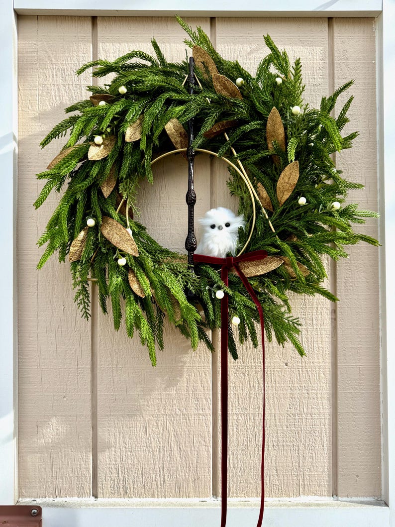 May include: A decorative wreath featuring green pine-like foliage, gold-toned leaves, and small white berries. A gold metal ring and a dark vertical rod are integrated into the design. A small, fluffy white owl figure sits inside, accented by a long dark red ribbon.