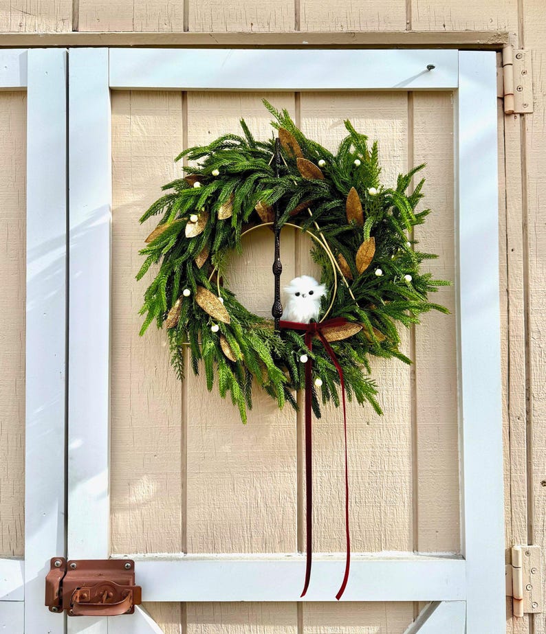 May include: A circular wreath made of green pine needles, accented with gold-toned leaves and small white berries. A small, fluffy white owl figurine sits in the center, framed by a gold metal ring and a dark red velvet ribbon hanging downwards.