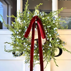 May include: A circular wreath made of green cedar branches and small, light blue berries. A long, deep red velvet ribbon is tied in a bow at the top, with two long tails hanging down the center of the wreath over the green foliage.