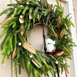 May include: A decorative wreath featuring green pine needles, gold-toned leaves, and small white berries. A black wand is positioned vertically in the center, accompanied by a small white fluffy owl figurine and a dark red velvet ribbon bow.