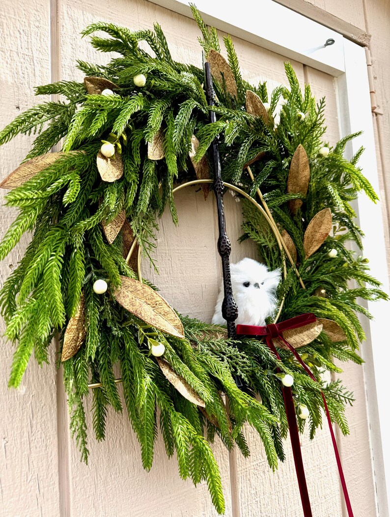 May include: A decorative wreath featuring green pine needles, gold-toned leaves, and small white berries. A black wand is positioned vertically in the center, accompanied by a small white fluffy owl figurine and a dark red velvet ribbon bow.