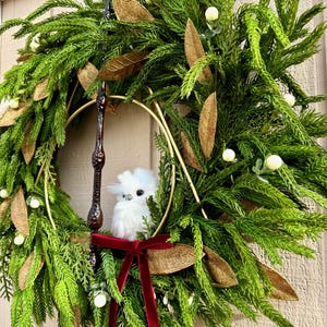 May include: A decorative wreath featuring green pine needles, gold-toned leaves, and small white berries. A gold metal triangle frame is centered within the wreath, holding a dark brown wand and a small, fluffy white owl figurine with a red velvet ribbon.