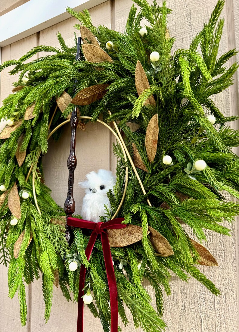 May include: A decorative wreath featuring green pine needles, gold-toned leaves, and small white berries. A gold metal triangle frame is centered within the wreath, holding a dark brown wand and a small, fluffy white owl figurine with a red velvet ribbon.