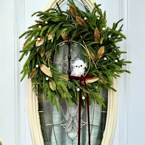 May include: A decorative wreath featuring green pine needles, gold-toned leaves, and small white berries. A dark brown wand is positioned vertically, intersecting a gold metal triangle. A small white plush owl sits in the center with a dark red velvet ribbon.