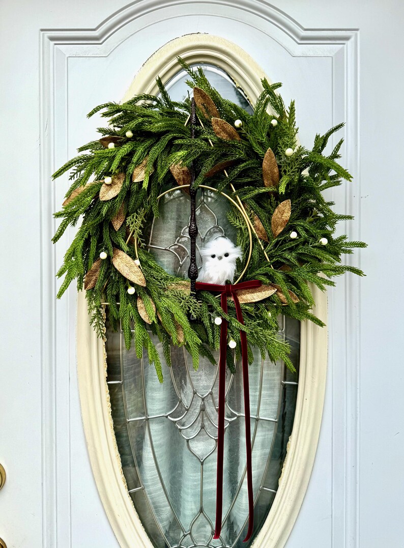 May include: A decorative wreath featuring green pine needles, gold-toned leaves, and small white berries. A dark brown wand is positioned vertically, intersecting a gold metal triangle. A small white plush owl sits in the center with a dark red velvet ribbon.