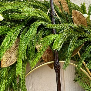 May include: A dark brown, textured wand with a knobby handle is positioned vertically against a decorative wreath. The wreath features vibrant green pine-like needles, metallic gold-toned leaves, and small white berries arranged on a gold metal ring.
