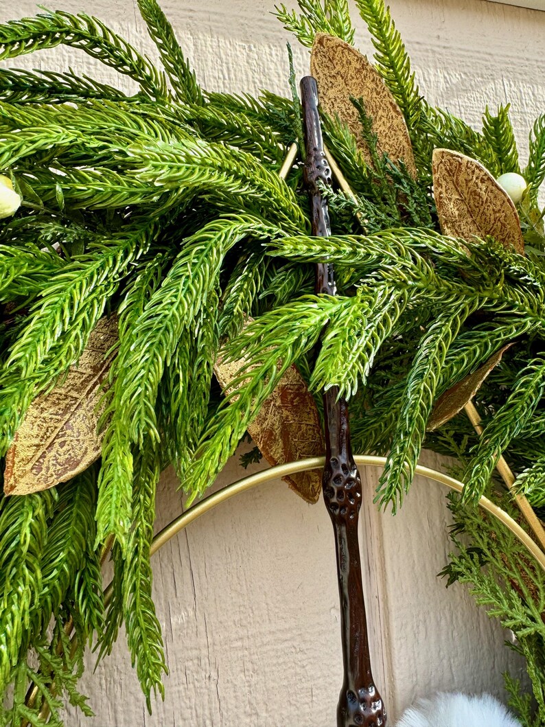 May include: A dark brown, textured wand with a knobby handle is positioned vertically against a decorative wreath. The wreath features vibrant green pine-like needles, metallic gold-toned leaves, and small white berries arranged on a gold metal ring.