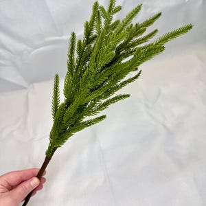 May include: A sprig of artificial greenery with vibrant green, needle-like leaves. The stem is a dark brown color. The sprig is held against a white background.