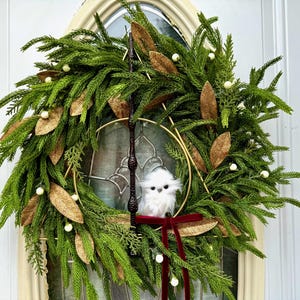 May include: A circular wreath made of green pine needles and cedar branches, decorated with gold-toned leaves and small white berries. A dark brown wand is positioned vertically, and a small white owl sits above a dark red velvet ribbon bow.