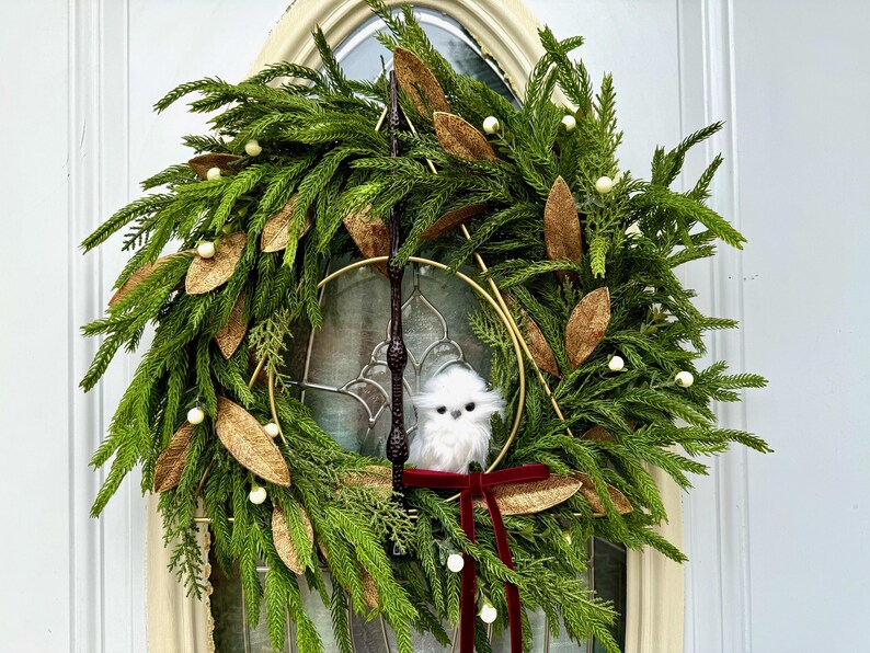 May include: A circular wreath made of green pine needles and cedar branches, decorated with gold-toned leaves and small white berries. A dark brown wand is positioned vertically, and a small white owl sits above a dark red velvet ribbon bow.
