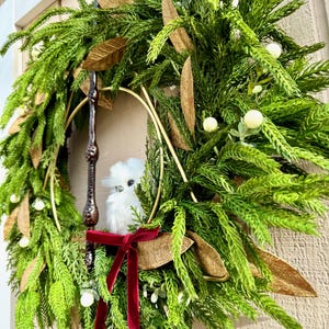 May include: A decorative wreath featuring green pine needles, gold-toned leaves, and small white berries. A dark brown wand is positioned vertically within the center, accompanied by a small white plush owl and a deep red velvet ribbon bow.