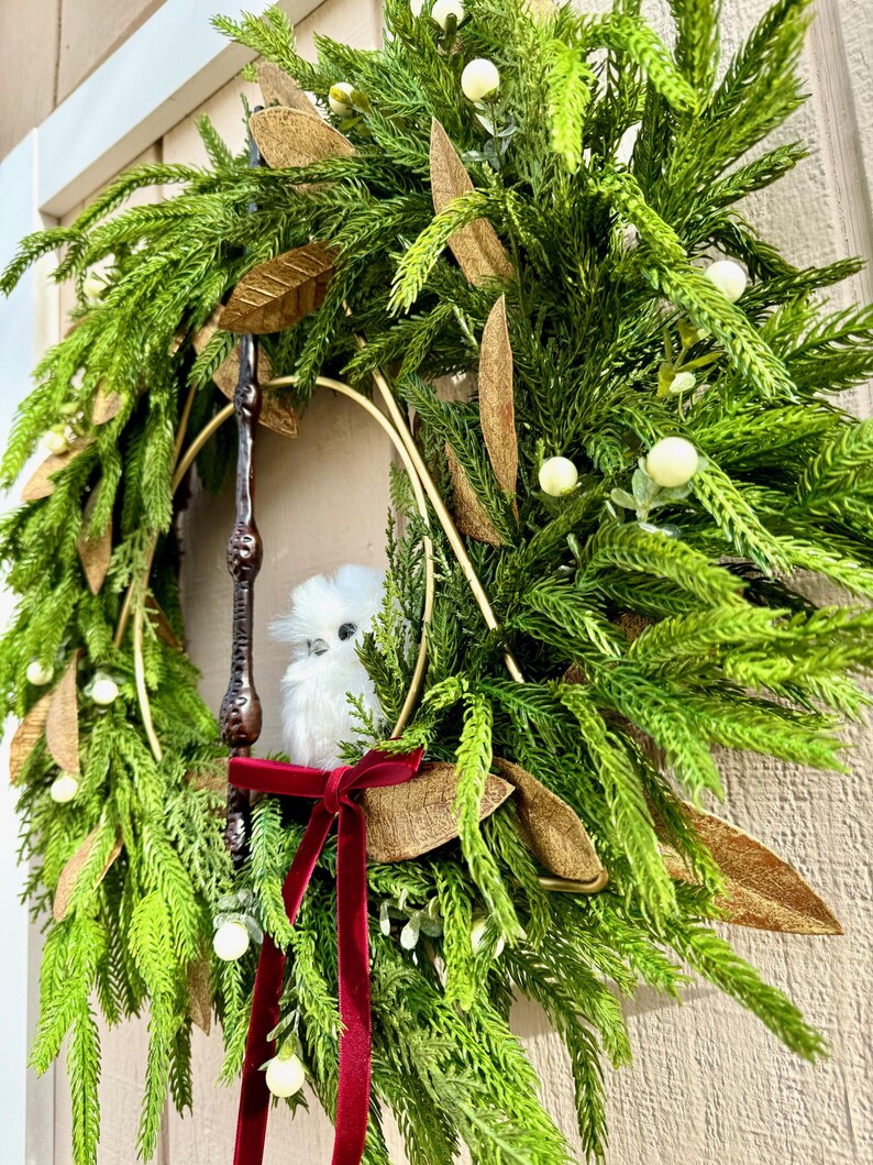 May include: A decorative wreath featuring green pine needles, gold-toned leaves, and small white berries. A dark brown wand is positioned vertically within the center, accompanied by a small white plush owl and a deep red velvet ribbon bow.