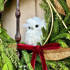 May include: A decorative wreath featuring green pine needles, gold-toned leaves, and a dark brown textured wand. A small, fluffy white owl figurine with black eyes sits in the center, accented by a deep red velvet ribbon tied in a bow.