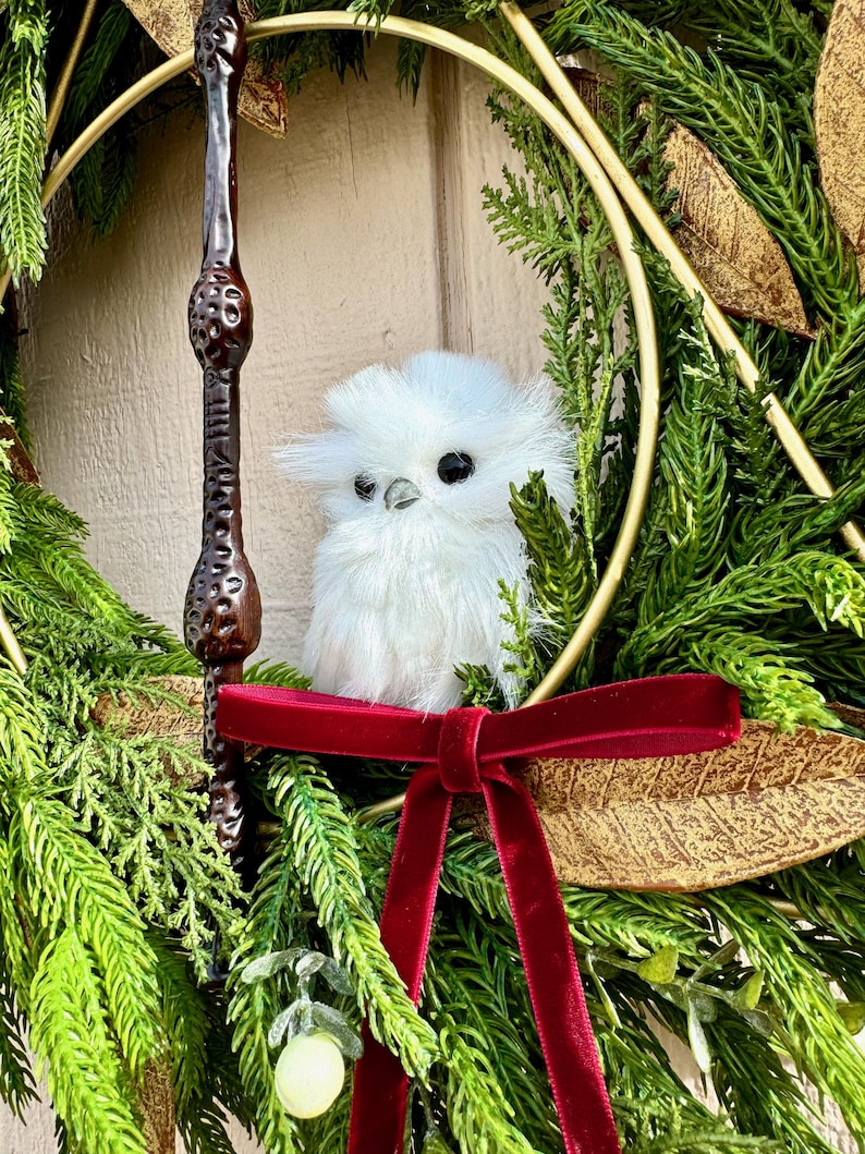 May include: A decorative wreath featuring green pine needles, gold-toned leaves, and a dark brown textured wand. A small, fluffy white owl figurine with black eyes sits in the center, accented by a deep red velvet ribbon tied in a bow.