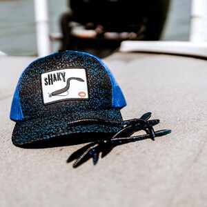 Peut inclure: Un chapeau de camionneur noir et bleu avec un patch blanc qui dit "SHAKY" et une illustration noir et blanc d'un leurre de pêche. Le chapeau est sur une surface grise avec un leurre de pêche bleu et noir.