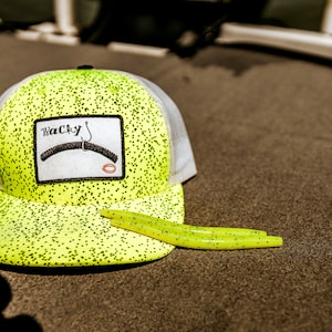 Peut inclure: Un chapeau de camionneur jaune et blanc avec un patch noir et blanc qui dit "Wacky" avec un hameçon et un ver de pêche. Le chapeau est posé sur une surface brune avec un leurre de pêche jaune et noir.