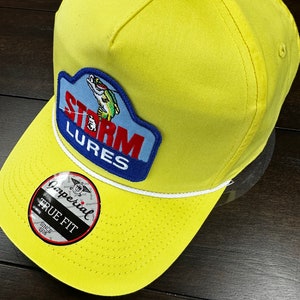 May include: Yellow baseball cap with a white rope trim and a blue and red patch that says "Storm Lures" with a fish graphic. The cap also has a black and white label that says "Imperial True Fit Since 1916."