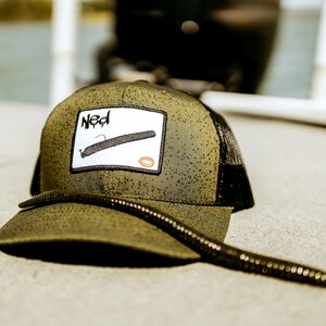 May include: A green and black trucker hat with a white patch that says "Ned" and a drawing of a fishing lure. A black worm is lying on the hat.