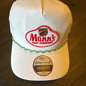 Può includere: Cappellino da baseball bianco con visiera curva. Il cappellino presenta il logo Mann's Bait Company in rosso e un profilo nativo americano. Una toppa circolare recita "Imperial True Fit Since 1916."
