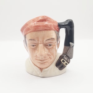 May include: A ceramic mug shaped like a man's head wearing a red hat. The mug has a black handle shaped like a leg and foot.