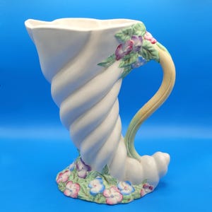 Puede incluir: Jarrón de cerámica blanca con diseño en espiral y asa curva. El jarrón está adornado con flores rosas, moradas y azules, y hojas verdes. Tiene una abertura ancha y una base en forma de concha.