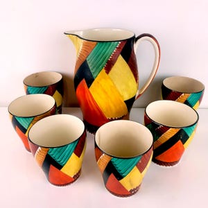 May include: A ceramic jug and six matching cups with a geometric pattern in red, yellow, green, and black. The jug has a curved handle and spout. The cups have a flared shape. The set is a vibrant addition to any table setting.
