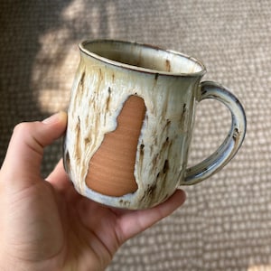 May include: A ceramic mug featuring a smooth, unglazed terracotta-colored patch on the side, contrasted by a glossy, cream-colored glaze with vertical brown drip patterns. The mug has a curved handle and a wide, rounded body shape.
