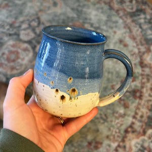 May include: A ceramic mug with a rounded body and a curved handle. The upper portion features a deep blue glaze, while the lower section is a creamy off-white. Small, dark brown speckles are visible where the two colors meet.