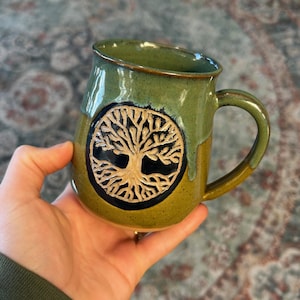 May include: A ceramic mug featuring a two-tone green glaze with a darker olive base and a lighter sage top. A circular medallion on the front displays a raised, light-colored relief of a tree with intricate branches and roots.