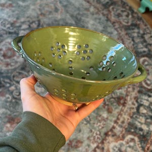 May include: A round, olive green ceramic colander featuring two side handles and a series of circular drainage holes distributed across the bowl. The interior and exterior surfaces have a glossy finish, with a visible unglazed tan base.