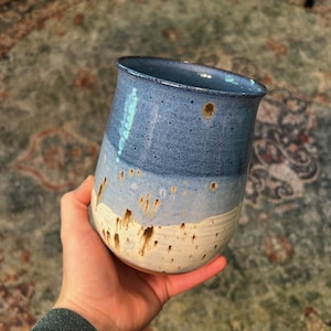May include: A ceramic tumbler featuring a gradient glaze that transitions from a deep blue at the rim to a lighter blue in the center and an off-white base. The surface is speckled with dark brown spots and has a smooth, curved silhouette.