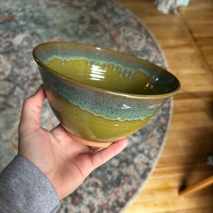 May include: Handmade ceramic bowl with a unique glaze. The bowl features a blend of colors, including a vibrant yellow-green base, a band of blue-green, and a brown rim. The bowl is held in a hand, showcasing its size and shape.
