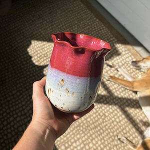 May include: A ceramic vessel with a ruffled rim, featuring a vibrant red upper section that transitions into a light blue and white base with brown speckles throughout the lower portion, showcasing a glossy finish and a rounded, bulbous shape.