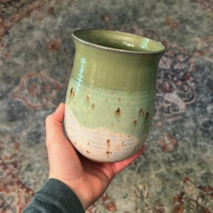 May include: A ceramic tumbler featuring a two-tone glaze with a smooth sage green upper section and a textured, speckled off-white base. The vessel has a slightly flared rim and a rounded body, showcasing a subtle drip pattern where the colors meet.