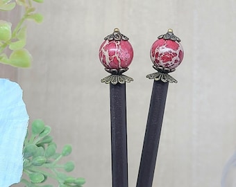 May include: Two black hair sticks with red and white marbled spherical tops. The tops are adorned with bronze-colored filigree and small gold accents. The hair accessories are displayed against a soft, neutral background.