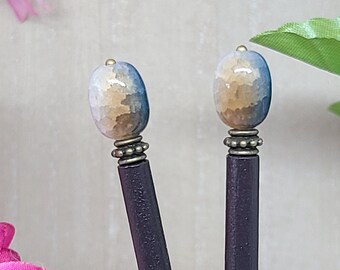 May include: Two decorative hair sticks. Each stick features a dark purple shaft, topped with a decorative bead in shades of blue, tan, and gold. The beads are accented with a brass-colored metal band.