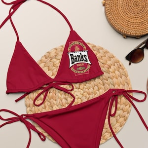 May include: Red bikini with a Banks Beer logo on the top. The bikini is on a woven straw mat with a round straw bag and sunglasses.
